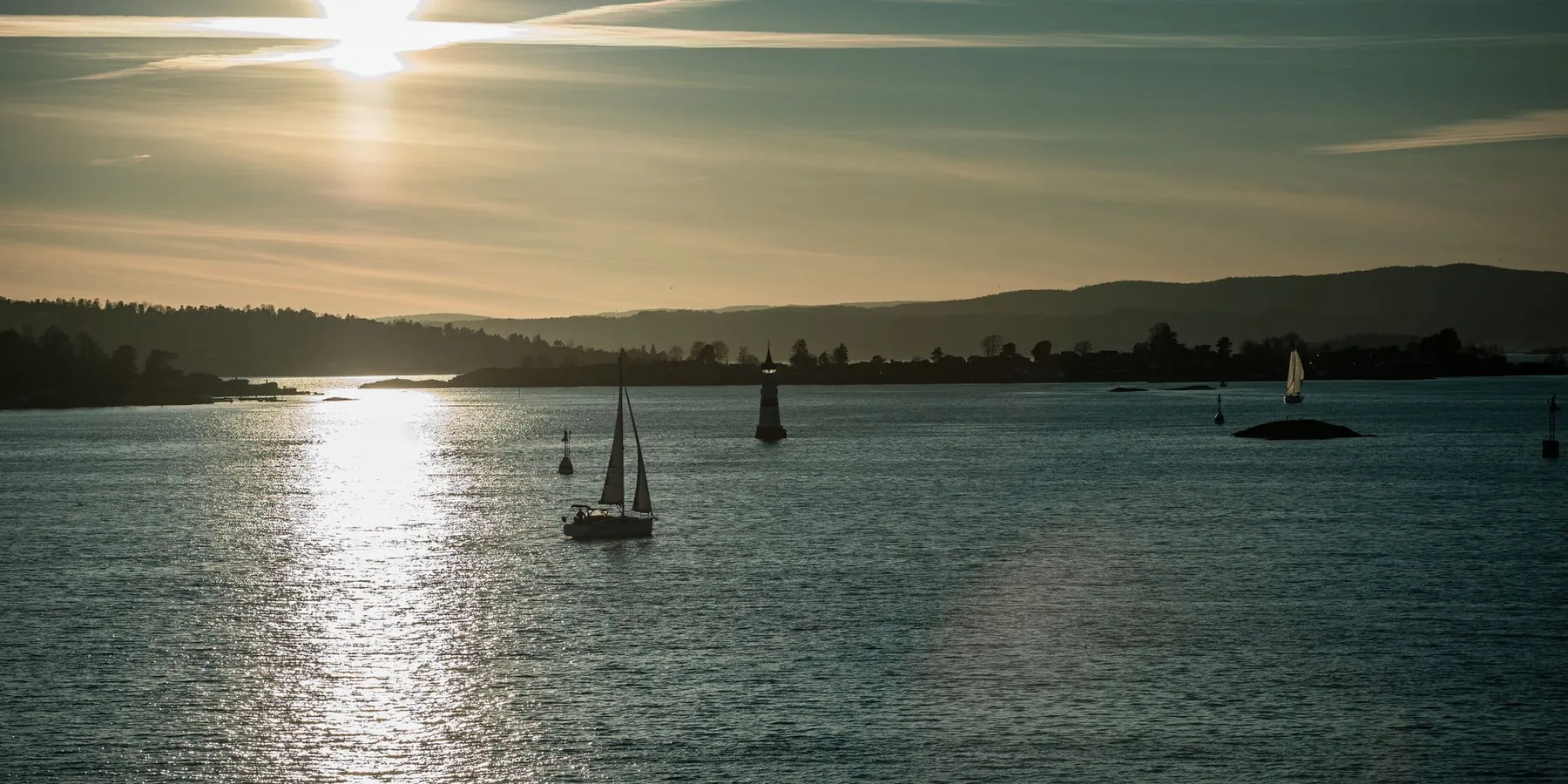 A sailing boat on the calm fjord with the sun low over wooded headlands