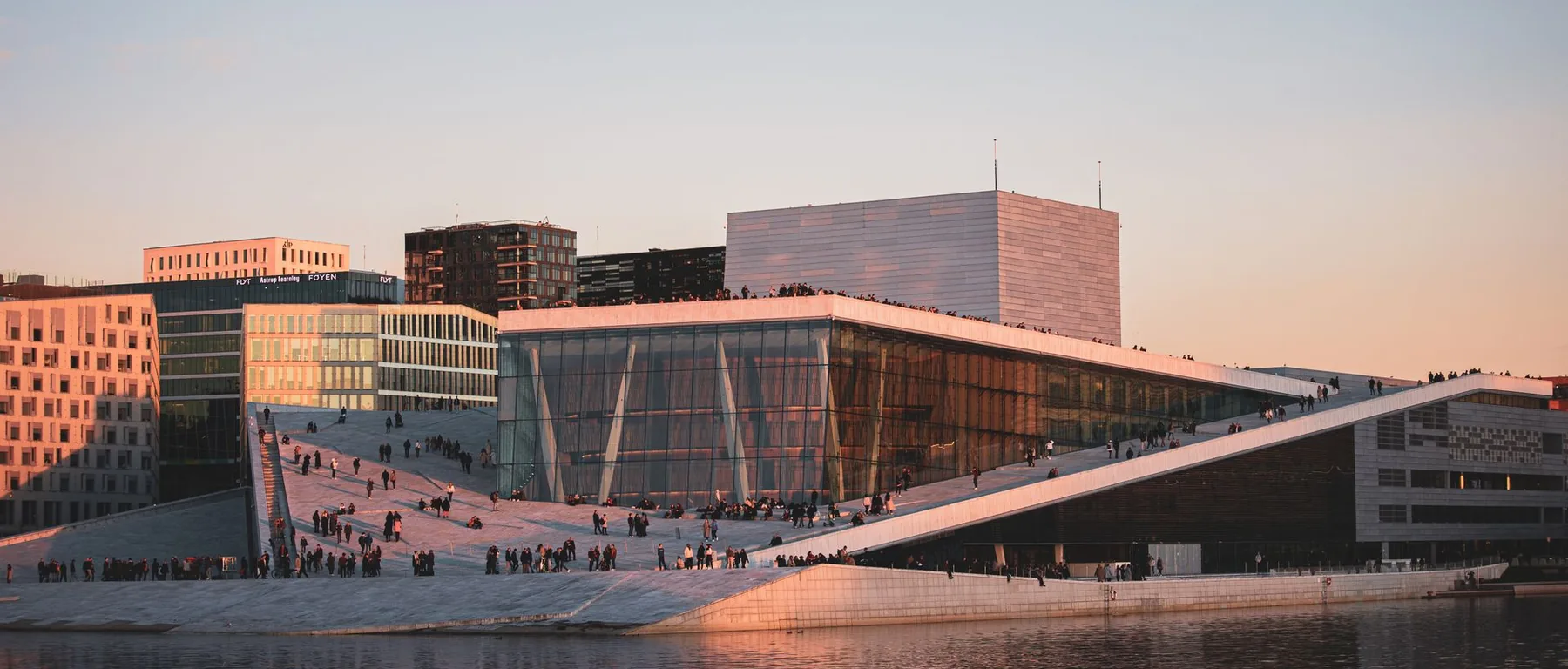 The Oslo Opera House at dusk, its white roof sloping to the water and catching the last warm light