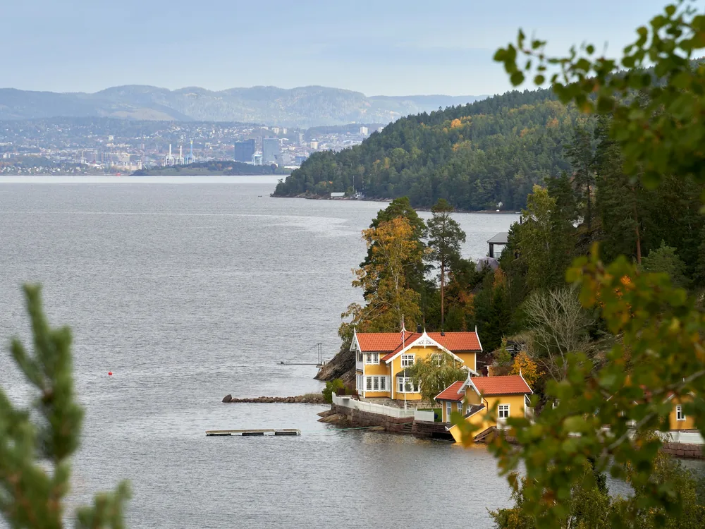 A yellow wooden house with a red roof on a wooded fjord shore