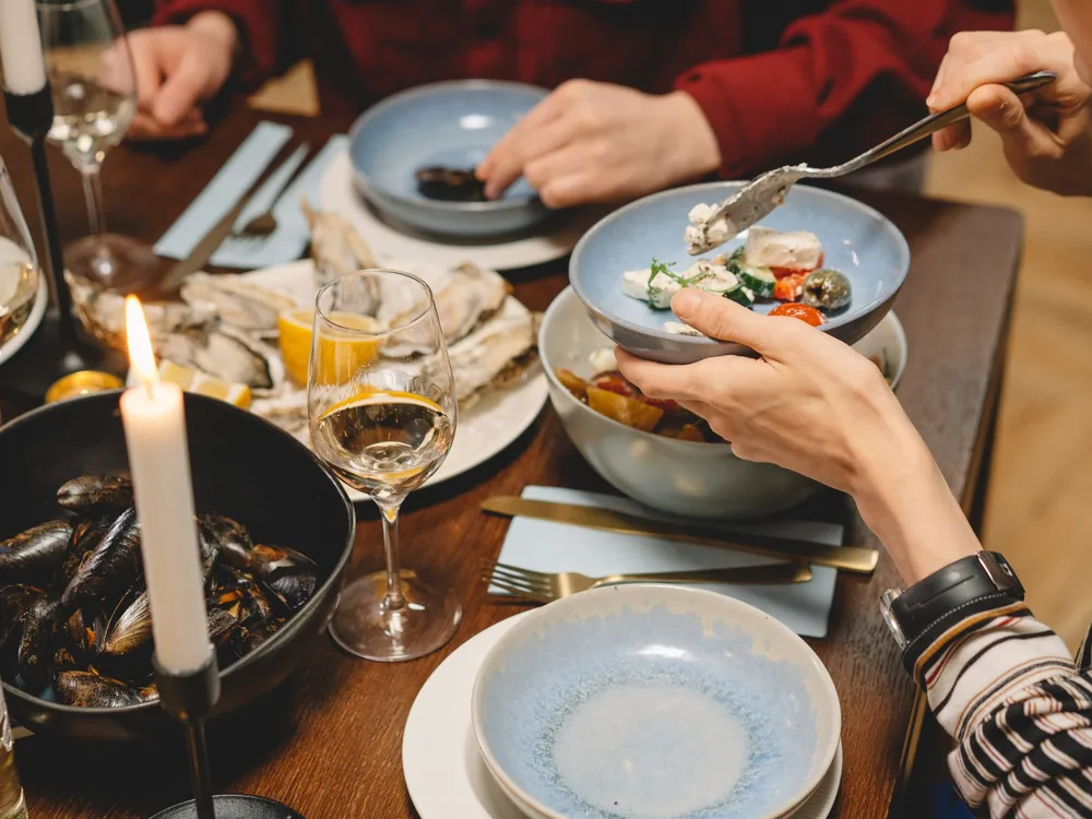 Hands sharing seafood dishes around a candlelit wooden table set with wine glasses